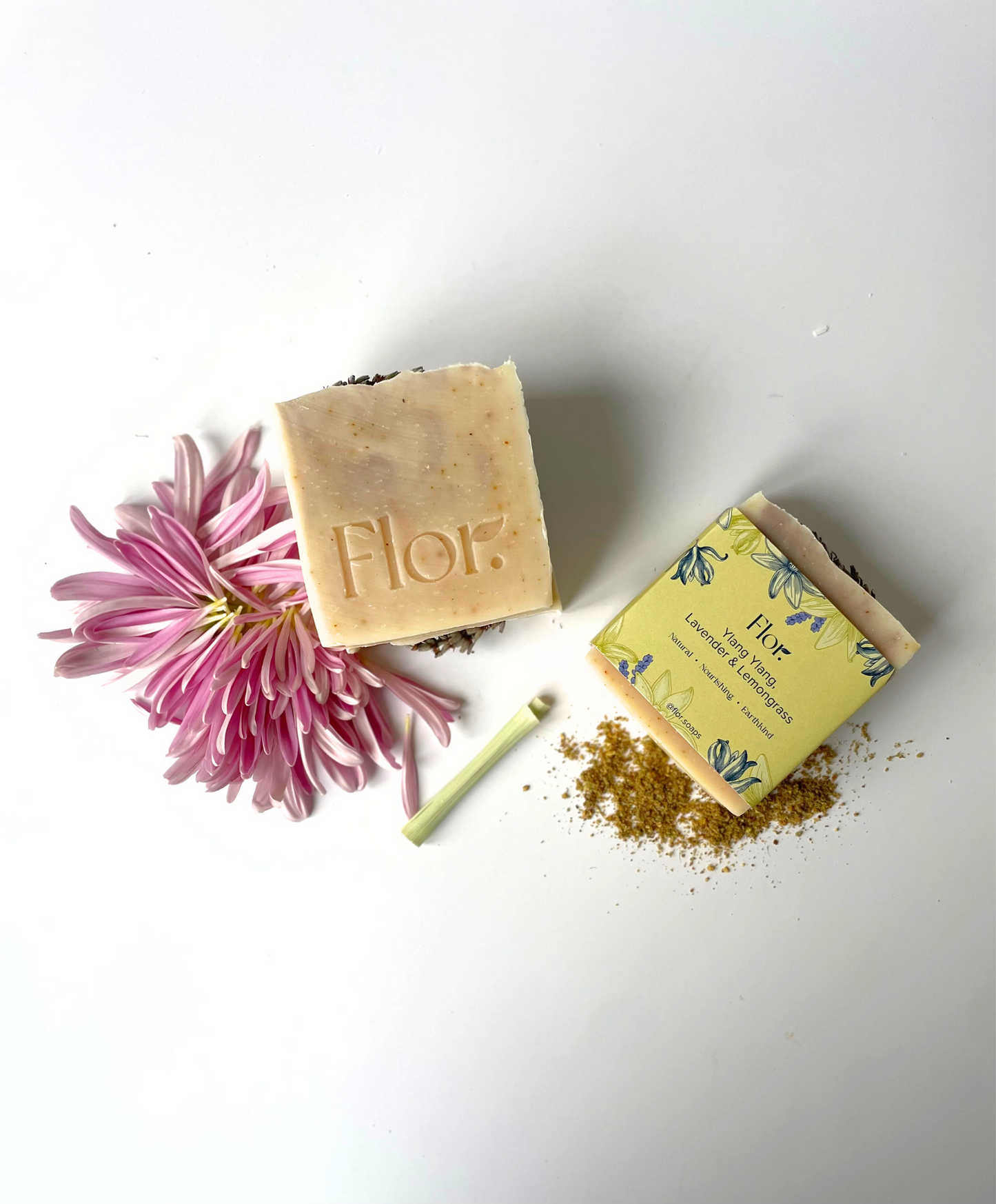 Two soap bars one with packaging with one without. Packaging is yellow and purple with flower illustrations and text information about the soap. The other bar has purple swirls from Brazilian clay and you can see the dried lemon peel and dried lavender flower in the soap and a dried lavender flower on top of the soap to decorate it