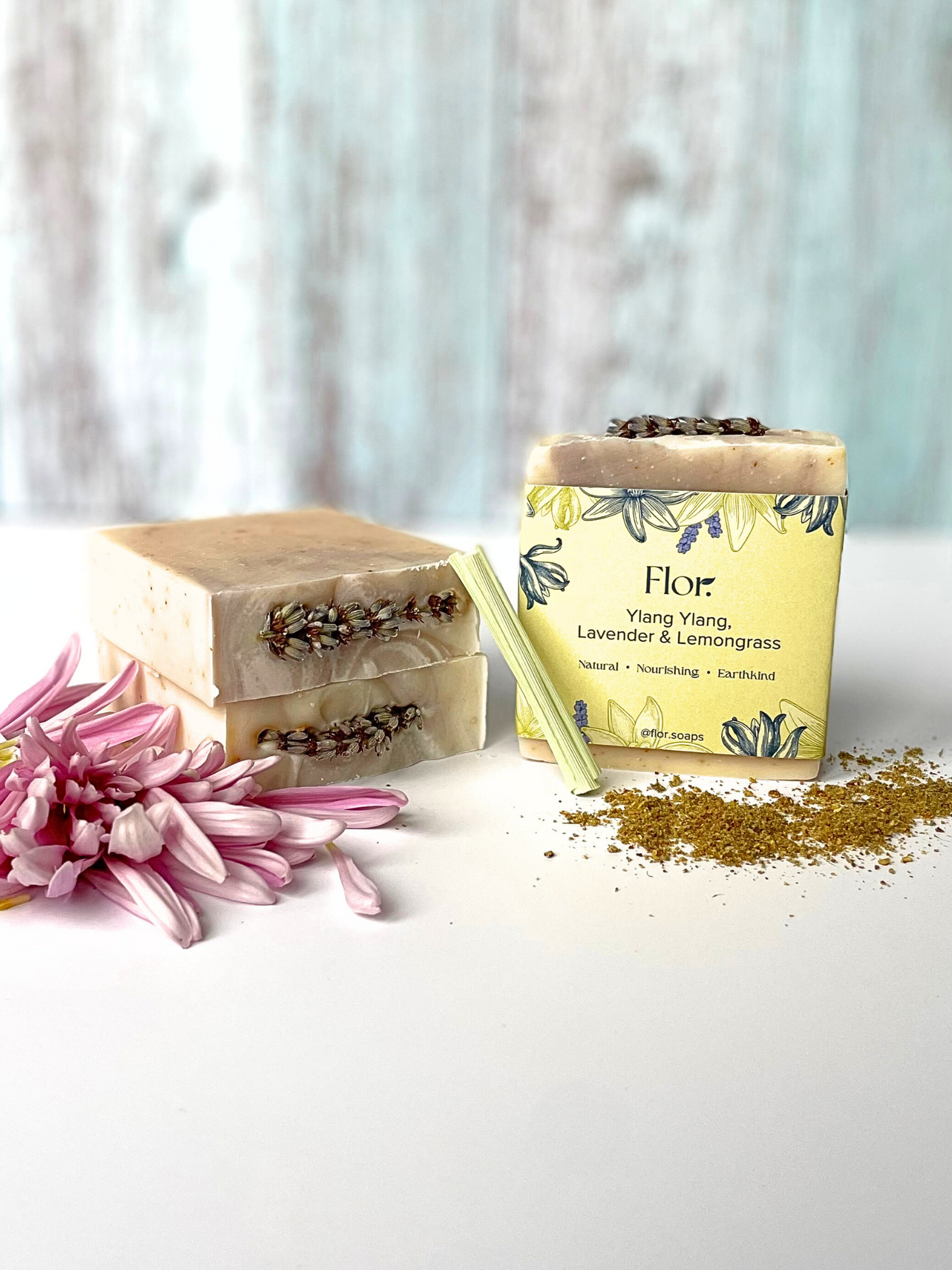 Three soap bars two laid on their side with dried lavender flower on the top the other has it packaging on which is yellow and purple with illustrations of flowers.
