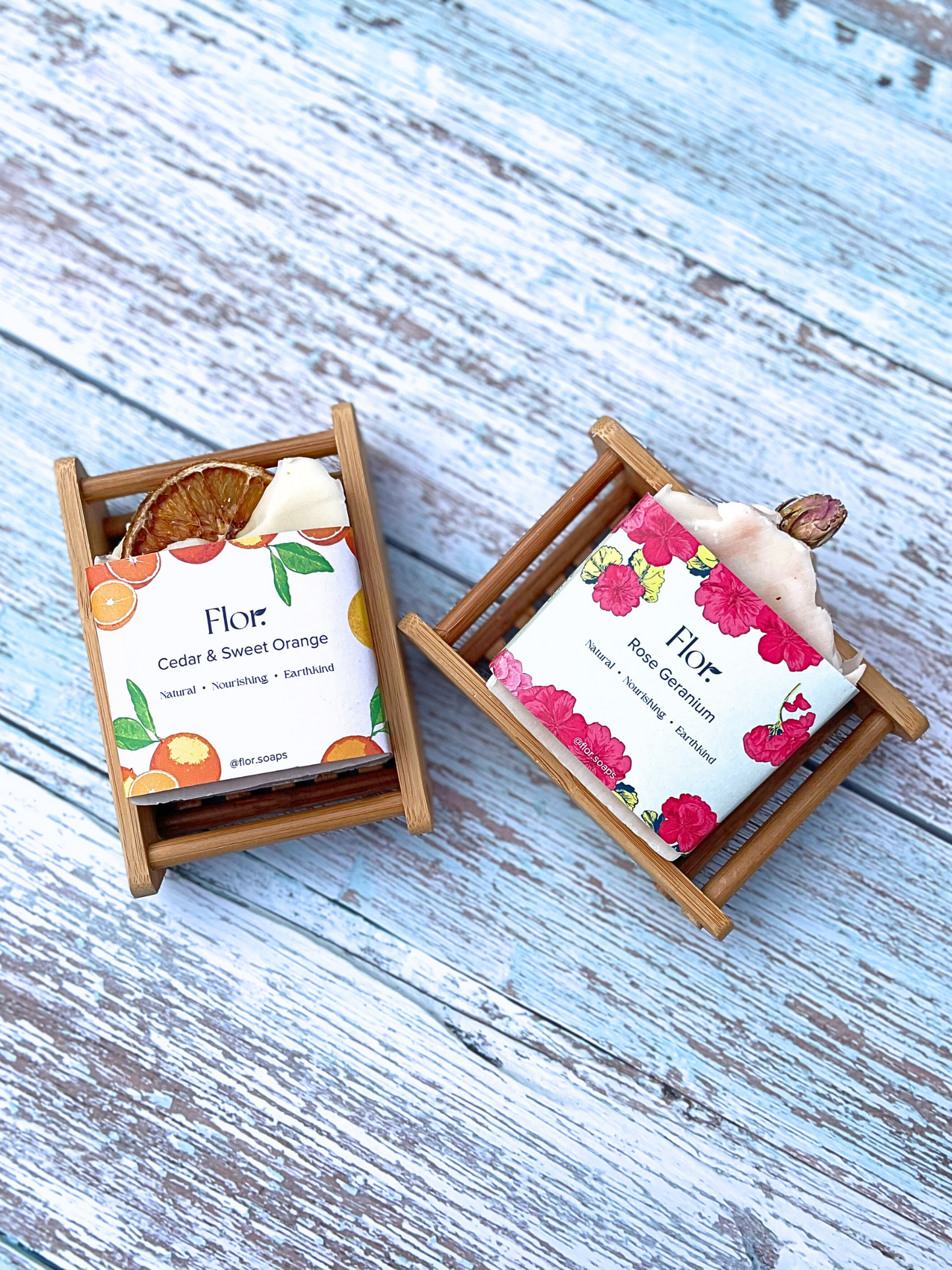 Two bamboo draining soap dishes displayed with two natural soap bars and their botanic packaging with floral and citrus illustrations