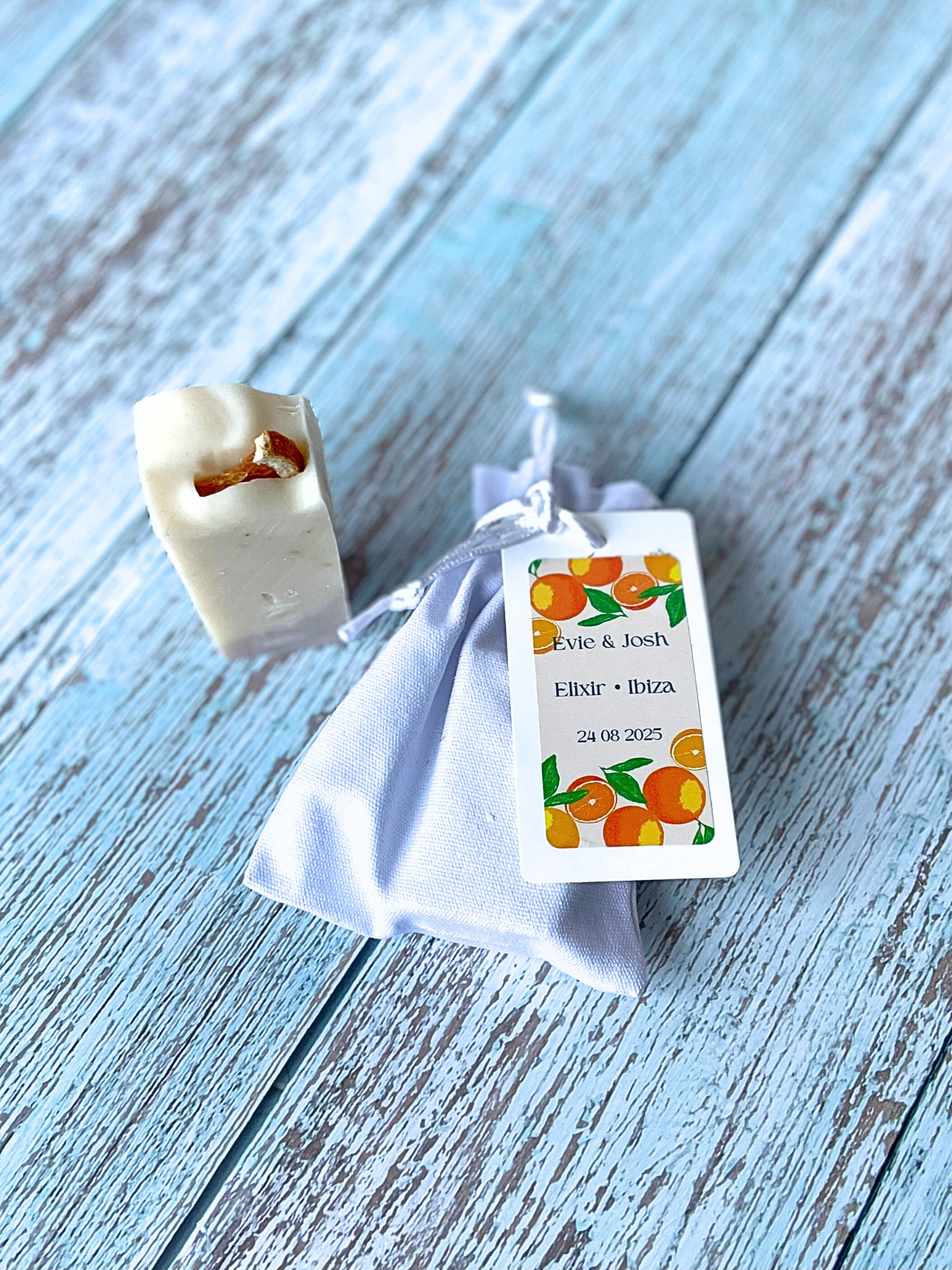 Mini soap favour is decorated with a dried orange slice, has a purple swirl made from Brazilian purple clay and is enriched with dried orange peel.  The image also includes a luxury white cotton bag with silk draw strings and colourful branding which is pink and orange with illustrations of oranges.  Can be used for weddings, baby showers, corporate events and corporate gifting