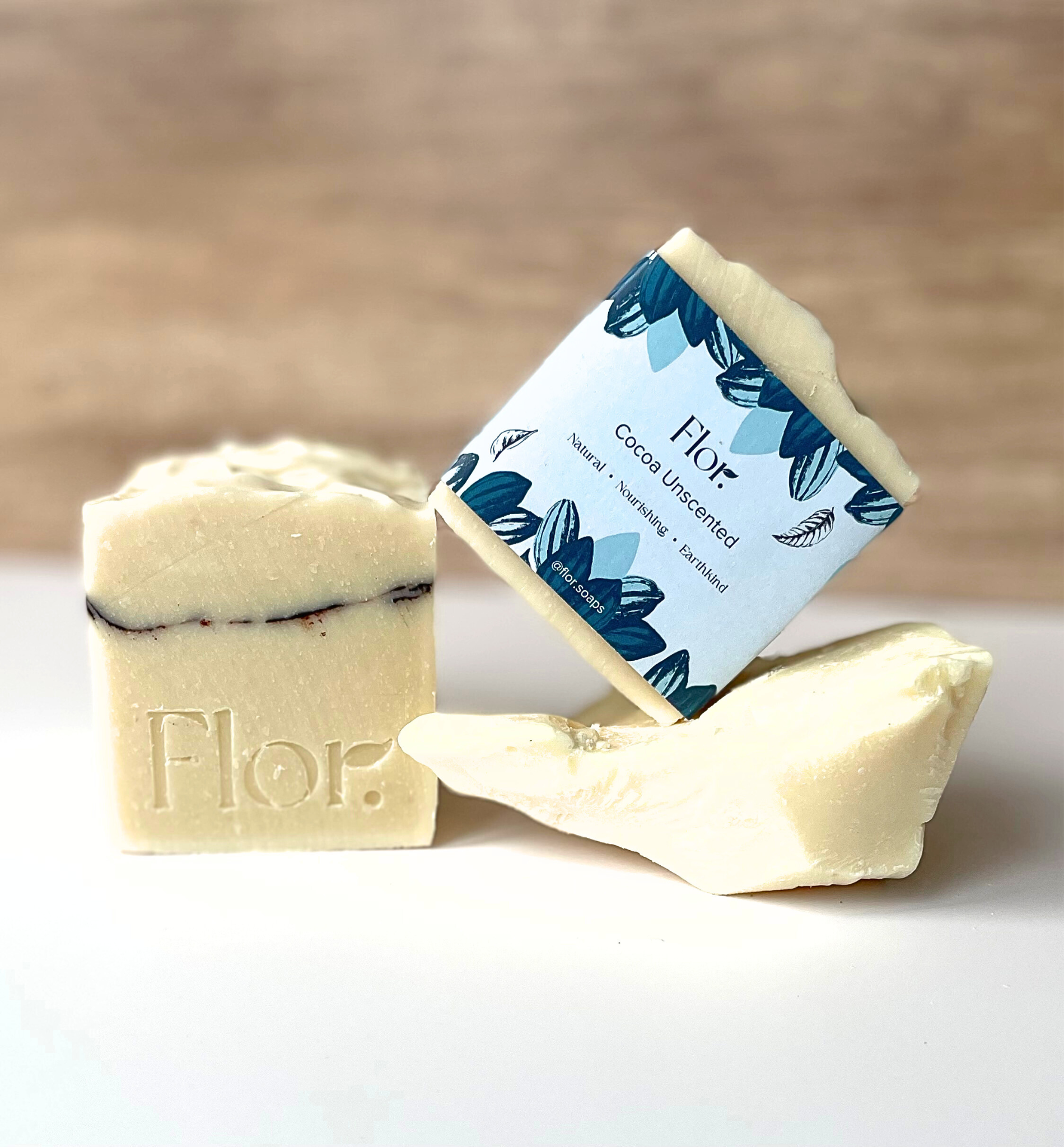 Cocoa unscented natural soap bar is displayed with its ingredients cocoa butter.  There are two soap bars in the image.  One is without packaging and has a decorative line going through it made of cocoa powder.  The other one has its packaging on which is pale blue background with darker blue illustrations of leaves and cocoa plants. 