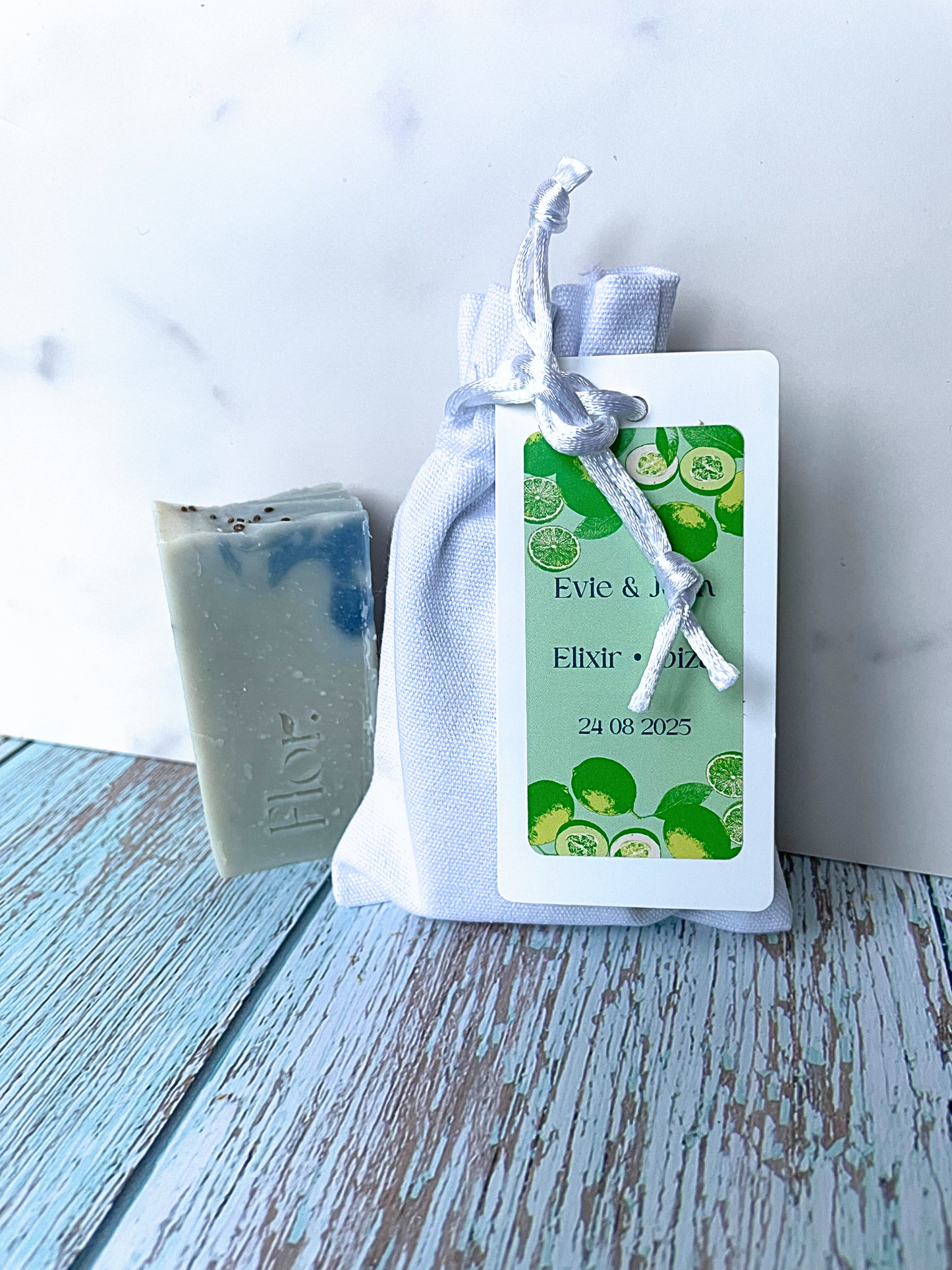 Cucumber spearmint and lime min natural soap favour for weddings baby showers corporate events ad corporate gifting.  The half bar soap favour is natural coloured blue with dark blue swirls. The image also includes the luxury white cotton bag with silk drawstrings that the soap comes in.  The tag on the bag has information on the soap and has colourful branding which is blue with green illustration of limes and cucumber.  Can be used with weddings baby showers corporate events and corporate gifting