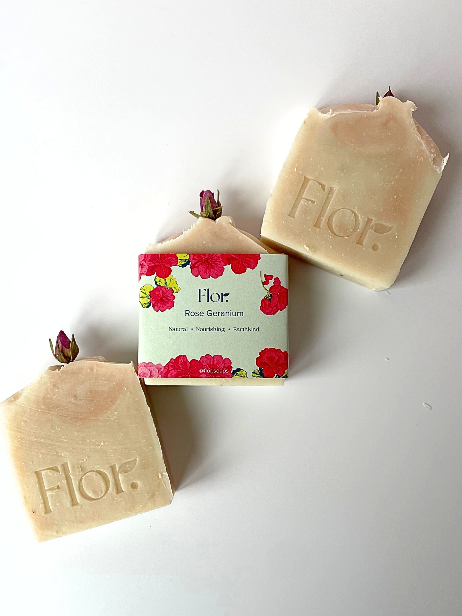 Three rose gernium natural soap bars are displayed and one has the packaging on it whih is bright and colourful with a green background nad pink rose geranium illustrations. The soap bars are decorted with a dried rose bud on the top and have light pink swirls in them made from French pink clay. 