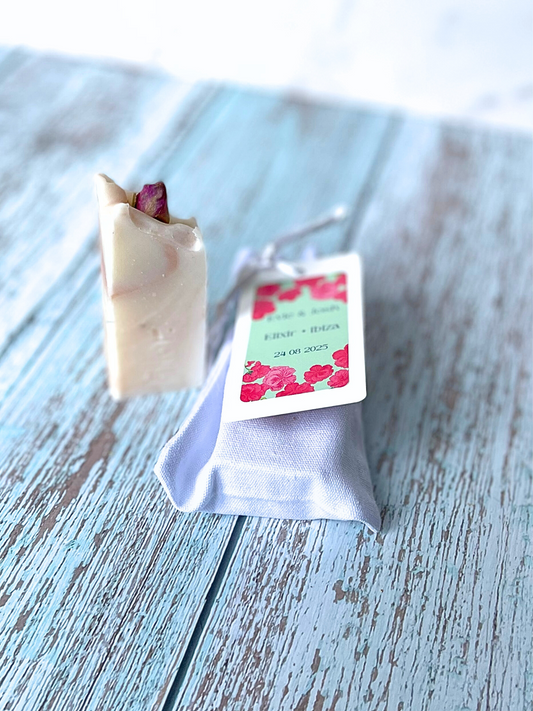 Wedding favour ideas.  Mini natural handmade soap bar used as a wedding or event favour decorated with a dried rose bud and has pink swirls in the bar from pink clay.  Included in the image is a luxury cotton bag with silk drawstring that the soap fits inside. The cotton bag has a tag with colourful branding and information on the soap bar.  This is green and pink with rose illustrations. Can be used as wedding favours, baby showers. corporate gifts, corporate gifting