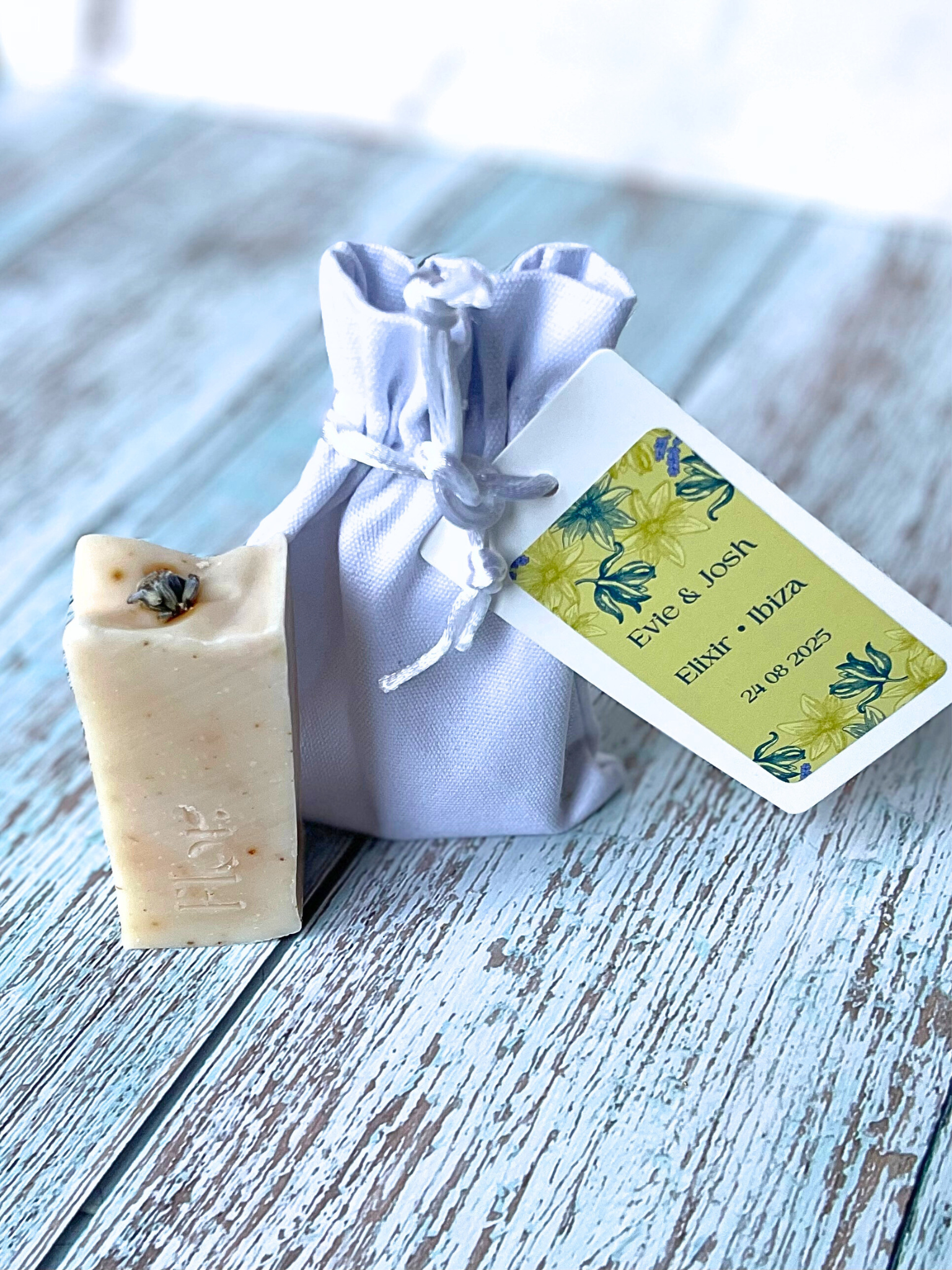 Mini soap favour with dried lavender flower decorating the top enriched with dried lemon peel and dried lavender flower.  Next to the soap bar is an image of the luxury cotton bag with silk draw strings that it comes in and it has a tag of colourful branding with the soaps information on it.  The tag is yellow and purple with purple flower illustrations.  Can be used for weddings baby showers corporate events and corporate gifting